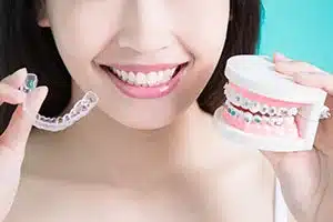 The bottom half of a smiling fgirl's face. She is holding a dental model with braces and a clear dental aligner.